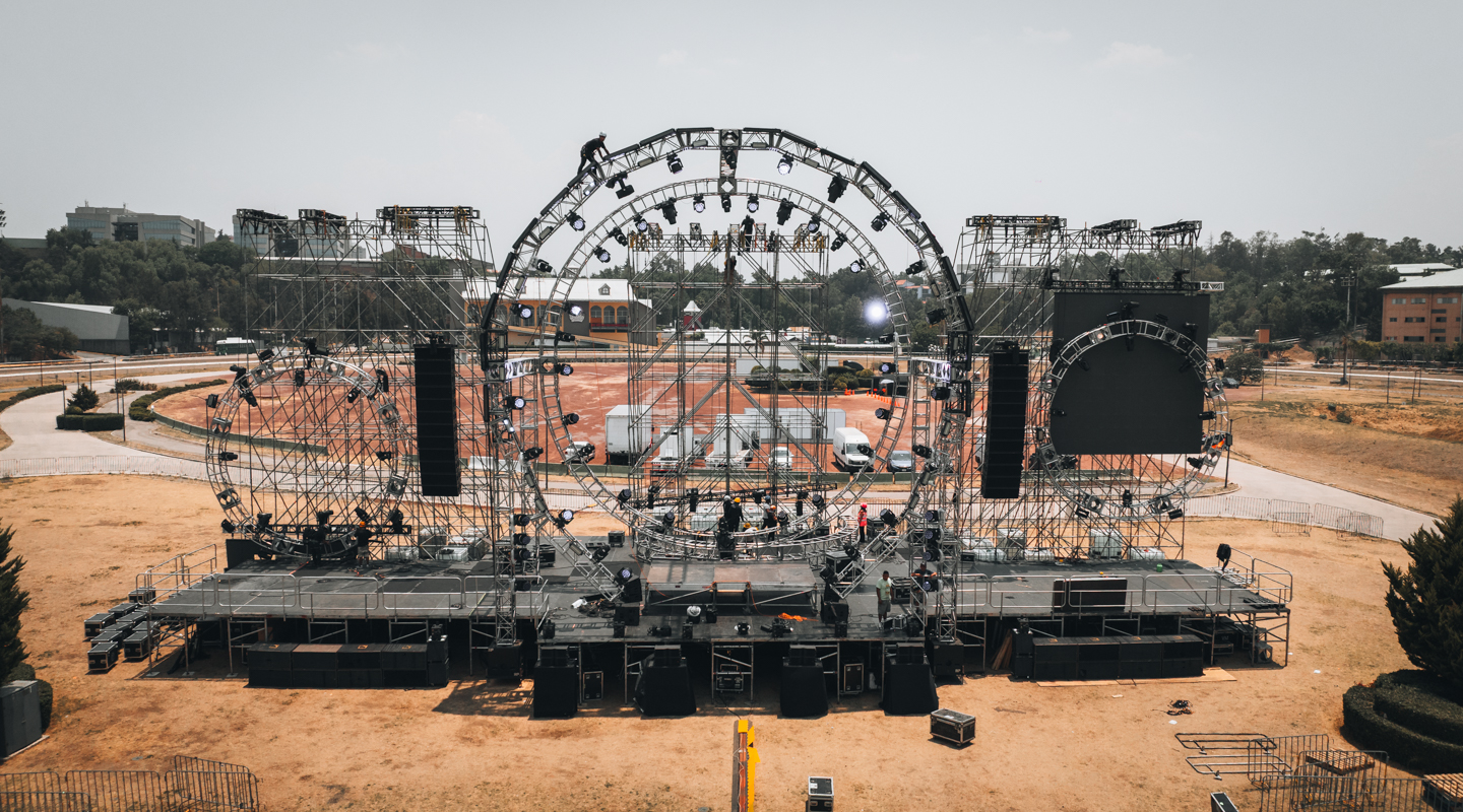 Vista lateral de unas gradas temporales con desgaste realista por uso, rodeadas de cables en el suelo de tierra y listas para recibir a los asistentes en un recinto de conciertos exterior.